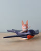 A toy rabbit named Spud the pilot Hare with a striped scarf, riding on Swifty Bird, a blue and red wooden bird figurine, set against a neutral beige background.
