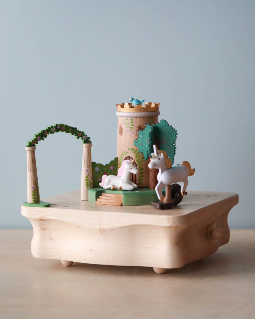The Unicorn Music Box is a hand-cranked music box with a small unicorn, white horse, castle tower, green archway, and trees on a light wood base against a pale blue background.