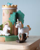 A close-up of the Unicorn Music Box shows a wooden unicorn with a gold mane and blue accents in front of a painted wooden castle and trees, with another unicorn in the background, creating a charming scene.