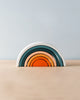 A minimalist image of a non-toxic, Handmade Mini Rainbow Stacker - Lagoon featuring arches in shades of teal, green, blue, orange, and red against a plain grey and sky blue background.