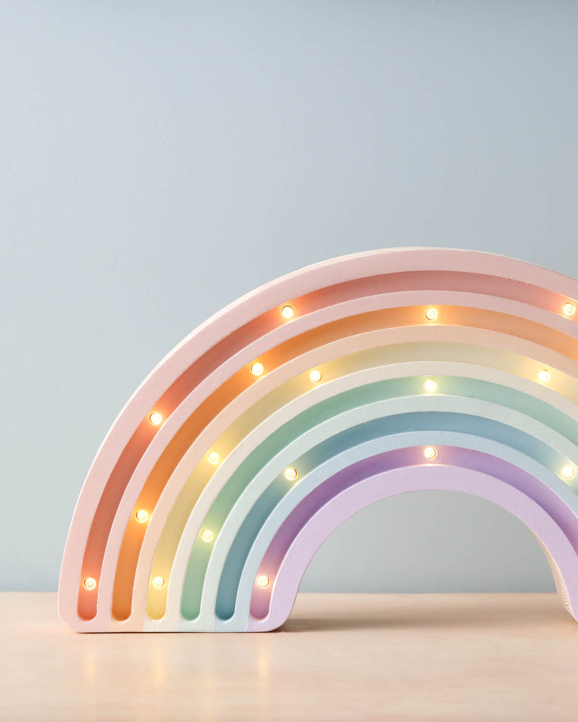 The Little Lights Rainbow Lamp, a pastel-colored children's night light with small bulbs illuminating each arc, sits on a pinewood surface against a light blue background.