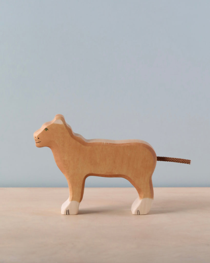 A handcrafted Holztiger Lioness wooden figurine, featuring white paws and a brown string tail, stands on a light surface with a pale blue background, highlighting its quality European craftsmanship.