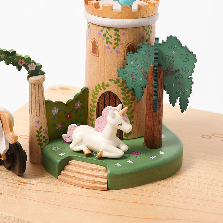 The Unicorn Music Box is a Wooderful Life hand-cranked scene with a white unicorn with a pink mane resting on green grass beside a tall castle tower, two trees, and floral decorations.