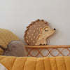 A cozy bedroom features the Little Lights Hedgehog Lamp with small illuminated spots, mounted on a white wall. Below is a woven rattan furniture piece with yellow and beige cushions, adding a warm and inviting atmosphere to the room. This adorable children's night light adds charm and comfort.