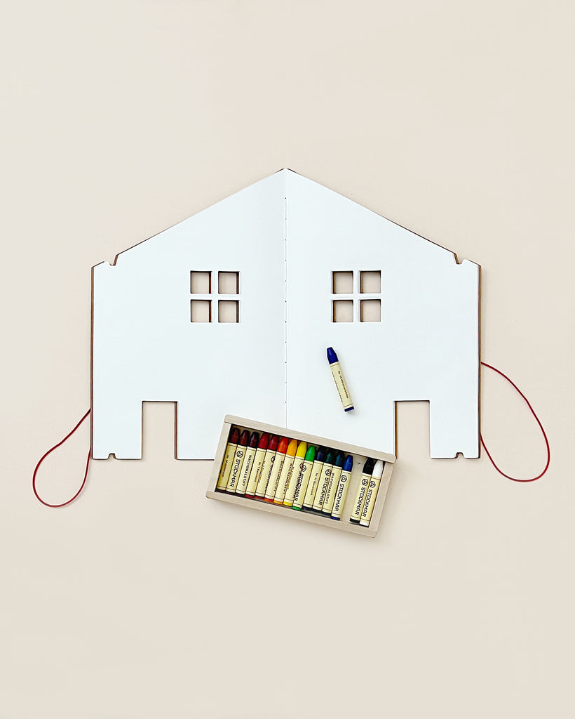 A creative flat lay featuring a white cardboard cutout of The Dollhouse Drawing Book split in half, with a red thread outlining its perimeter, and a box containing colorful thread spools placed beneath it, alongside a drawing