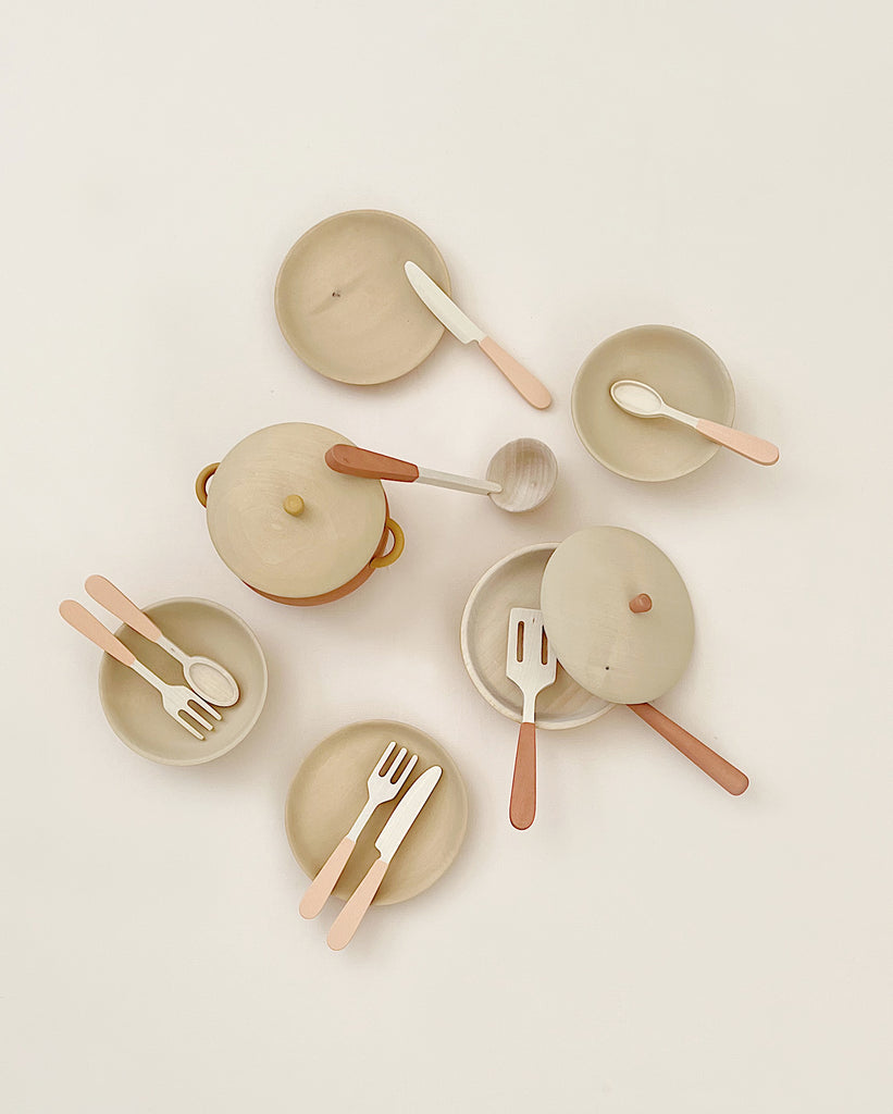 A minimalistic flat lay of a Handmade Wooden Kitchen Essentials - Flower, featuring wooden play kitchenware like bowls, a pot with a lid, plates, spoons, forks, and a spatula. The utensils and dishes are arranged in a circular pattern on a white background. Made with non-toxic paint for safe play.