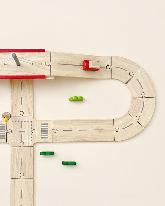 Wooden Road System Deluxe shaped like the letter 'd' with a red and yellow train car, and scattered green pieces on a light background, crafted from reclaimed rubberwood.