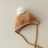A soft brown Briar Baby Saddle Pom Bonnet with ear flaps and a white pom-pom on top, laid flat on a light beige background.