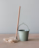 A simple, small green bucket with a metal handle next to a Maileg Miniature Mop & Bucket on a wooden surface against a neutral background, perfect for MINI sized Maileg friends.