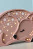 A Little Lights Deer Lamp, this pink wooden children's night light is shaped like a sleeping fawn with small warm white LED lights forming spots on its back. Handmade from pinewood, it highlights features like closed eyes and ears as the fawn rests against a light blue background.