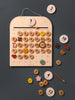 A My Calendar against a gray background, featuring magnetic discs with numbers and symbols, displayed on a board shaped like a house with weekdays labeled at the top.