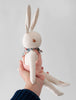 A person holds a Polka Dot Club Cream Rabbit in Hand Knit Overalls dressed in baby alpaca overalls and a floral scarf, against a plain light background. The rabbit has long ears, hand embroidered eyes, and dangling limbs.