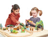 Two young girls playing with a Wild Pines Train Set that includes tracks, trees, buildings, and toy vehicles, interacting happily over their imaginative play setup.