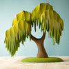 A handmade Large Wooden Willow Tree with cascading green foliage stands on a light-colored wooden surface against a pale blue background. The children's tree toy has a whimsical, stylized appearance with leaves that resemble fringed layers, all painted with non-toxic paint.