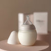 A modern Medical Grade Silicone baby bottle and its lid on a round pink table, with a soft beige background and a boxed product visible behind it. The bottle features a silicone nipple and a minimalist design.