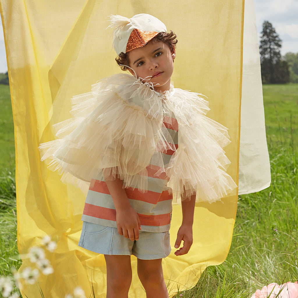 A young child in a playful Meri Meri Chick Costume - Final Sale featuring a striped shirt and tulle accessory, stands in front of a flowing yellow fabric backdrop in a sunny meadow.
