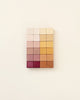 A grid of colorful square wooden blocks arranged in a gradient from white to deep red, coated with non-toxic paint, mounted on a light beige wall.