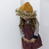 A young girl in a playful pose, wearing The Blueberry Hill Lion Hat and a mustard scarf, with her face partially obscured, against a white background.
